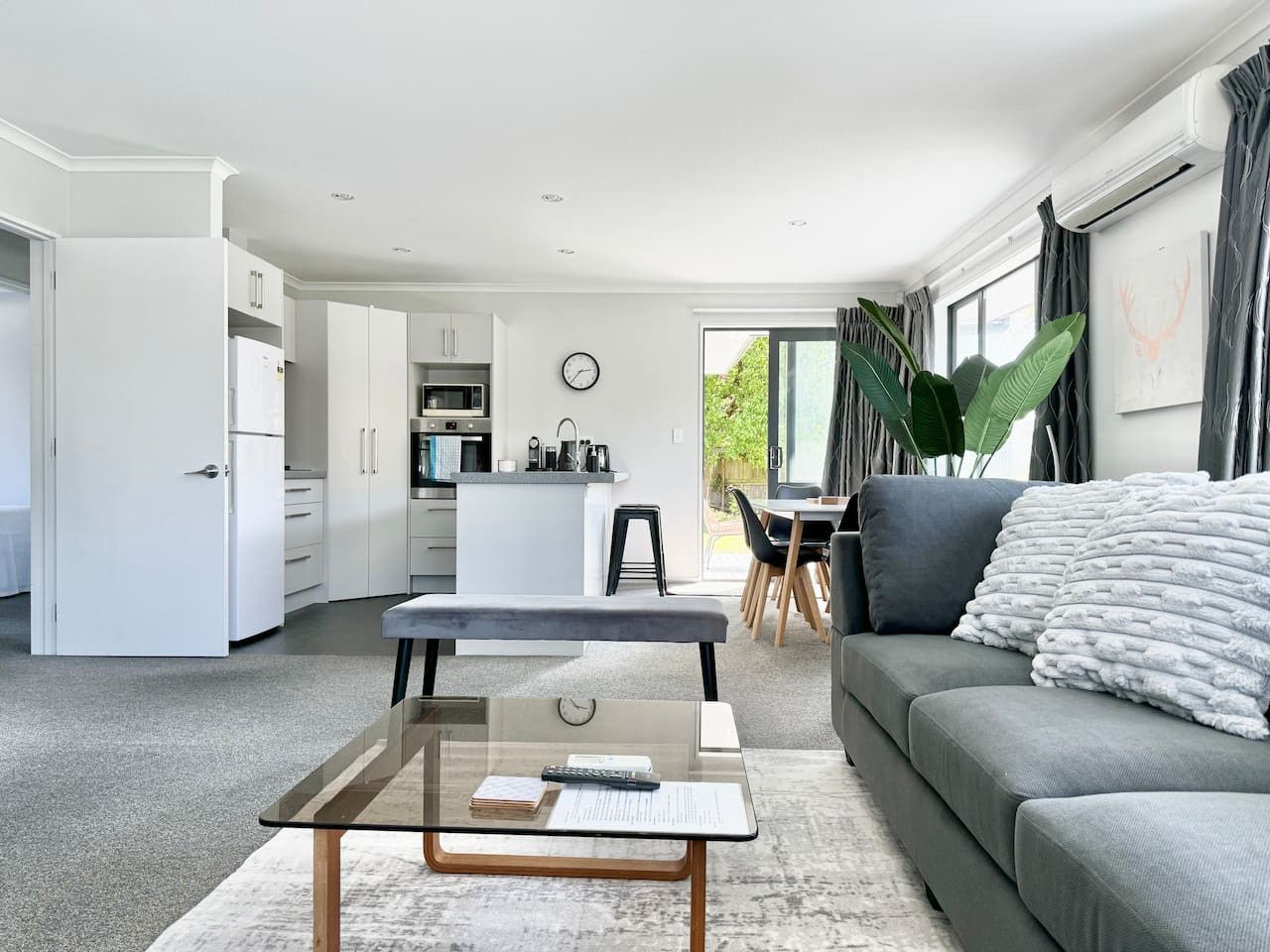 A modern living room with a glass coffee table, grey sofa, and a dining area with a plant.