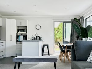 A modern kitchen with white cabinets, a built-in oven, and a sink. There is a dining area with a table and chairs, and a large window providing a view of the outdoors.