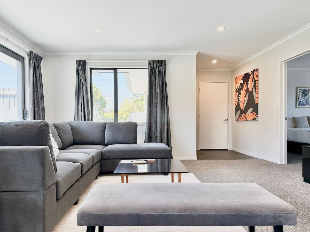 A modern living room with a grey sectional sofa, a small black coffee table, and a grey ottoman. There is a large window with grey curtains, a door, and a framed artwork on the wall.