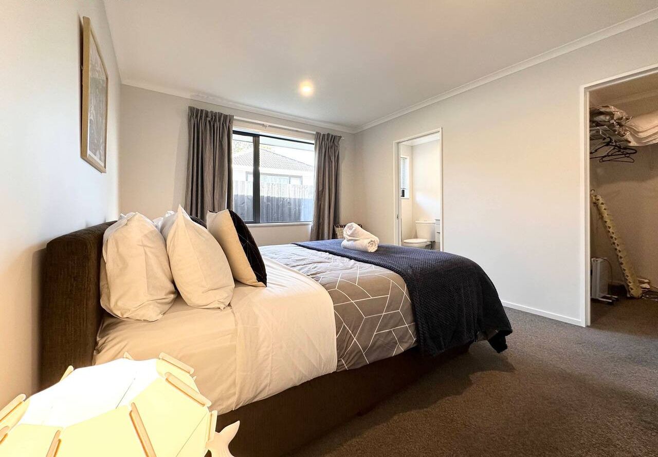 A master bedroom with a large bed, a window with curtains, a bedside table with a lamp, and a closet with hanging clothes.