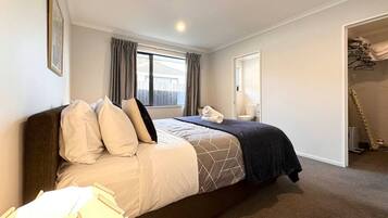 A master bedroom with a large bed, a window with curtains, a bedside table with a lamp, and a closet with hanging clothes.