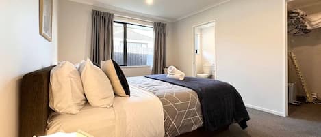 A master bedroom with a large bed, a window with curtains, a bedside table with a lamp, and a closet with hanging clothes.