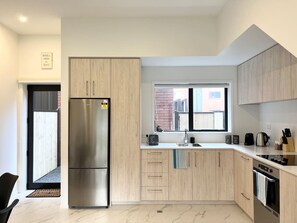 A modern kitchen with light wood cabinets, a stainless steel refrigerator, a built-in oven, and a window providing natural light.
