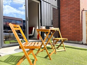 An outdoor seating area with wooden chairs and a table, a glass door entrance, and a brick wall. - Cashel townhouse with courtyard and baby gear in Christchurch (Christchurch)
