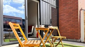 Un espace extérieur aménagé avec des chaises et une table en bois, une porte d’entrée en verre et un mur en briques.