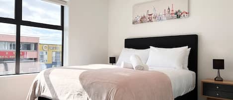 A bedroom with a large bed, a nightstand, a lamp, and a window with a view of buildings.