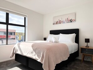 A bedroom with a large bed, a nightstand, a lamp, and a window with a view of buildings. - Cashel townhouse with courtyard and baby gear in Christchurch (Christchurch)