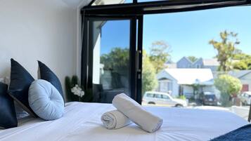 A neatly made bed with white linens and rolled towels, a dark blue pillow, and a view of the street and houses through a large window.