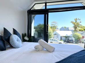 A neatly made bed with white linens and rolled towels, a dark blue pillow, and a view of the street and houses through a large window. - Peacock townhouse with garage and balcony in Christchurch (Christchurch)