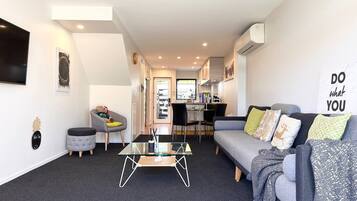 A modern living room with a glass coffee table, grey sofa, and a dining area with a kitchen in the background.