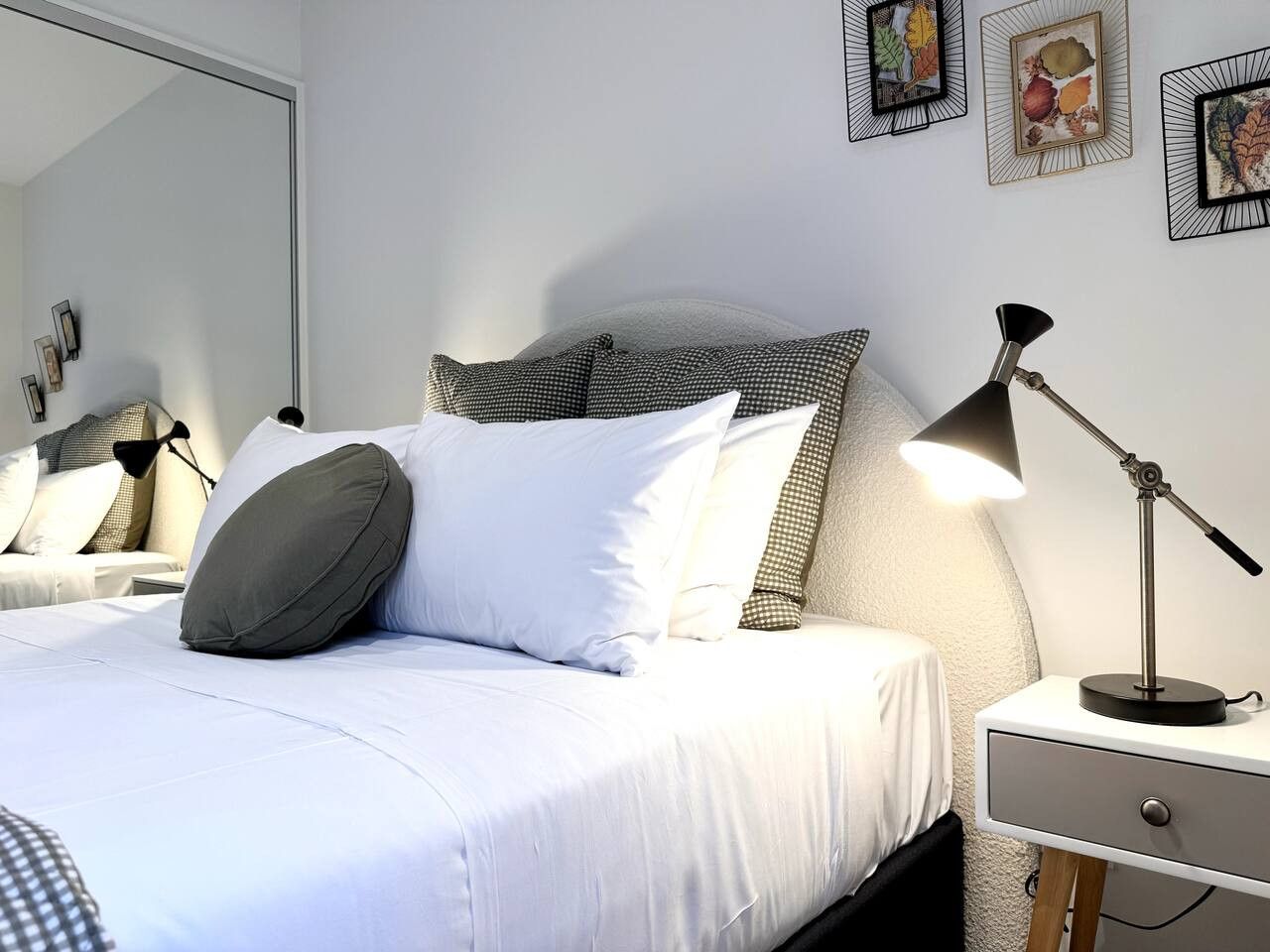 Een netjes opgemaakt bed met witte lakens en meerdere kussens, een nachtkastje met een lamp en lijstjes met kunst op de muur.