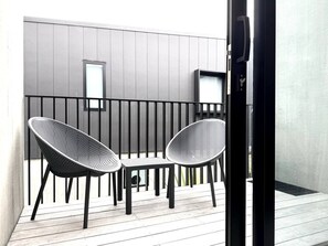 Ein Balkon mit zwei gewobenen Stühlen, einem kleinen Tisch, einer Schiebetür und einer grauen Wand mit vertikalen Feldern.
