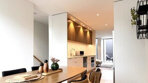 A modern dining area with a wooden table and chairs, a kitchen in the background, and a view of the outdoors.