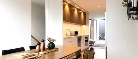 Un espace repas moderne avec une table et des chaises en bois, une cuisine en arrière-plan et une vue sur l’extérieur.
