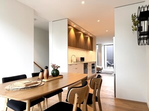 Un comedor moderno con una mesa y sillas de madera, una cocina al fondo y vistas al exterior.