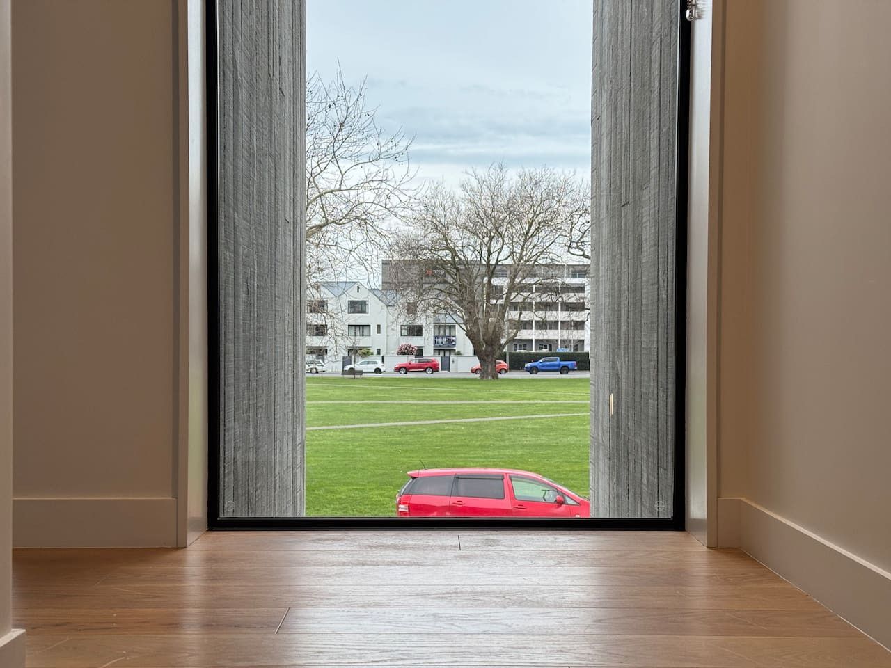 A room with a wooden floor and a view through a glass door to a grassy area with a red car and buildings in the background.