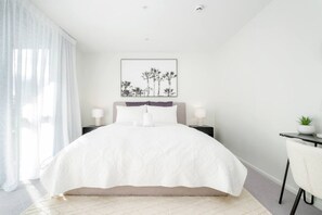 Une chambre moderne avec un grand lit, deux tables de chevet avec des lampes, un tableau encadré au mur et une petite table avec une plante.