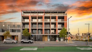 A modern multi-story building with a green bike lane in front, two cars parked on the street, and a sunset sky. - Cashel penthouse with city balcony and parking in Christchurch (Christchurch)