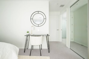 Ein modernes Schlafzimmer mit Bett, Schreibtisch mit Stuhl, einer kleinen Pflanze und einem runden Spiegel an der Wand.