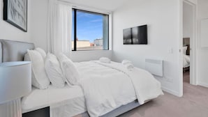 Quarto moderno com cama grande, mesa de cabeceira, janela com vista para a cidade e televisão na parede.