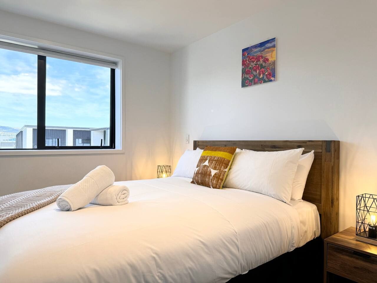A neatly made bed with white linens and a wooden headboard, a window with a view of buildings, and a painting on the wall.