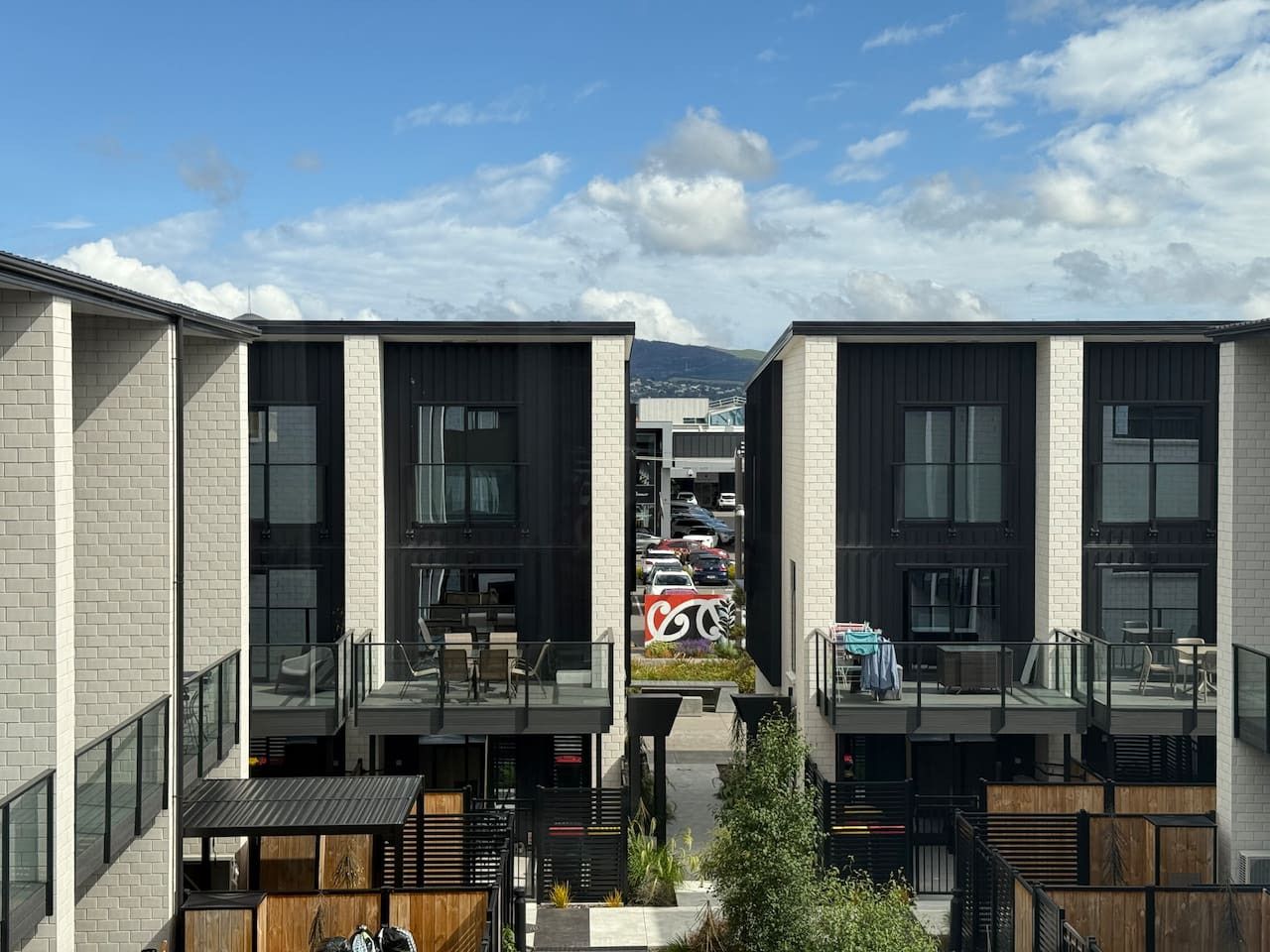 A modern building with two-story balconies, large windows, and a view of parked cars and mountains in the background.