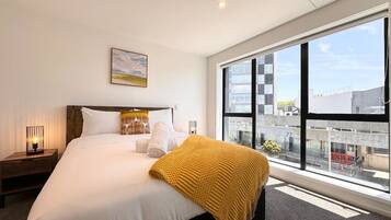 A hotel room with a large bed, a window with a city view, a bedside lamp, and a framed picture on the wall.