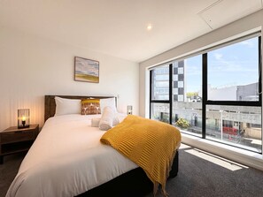 Una camera d'albergo con un letto grande, una finestra vista città, una lampada da comodino e un quadro appeso al muro.