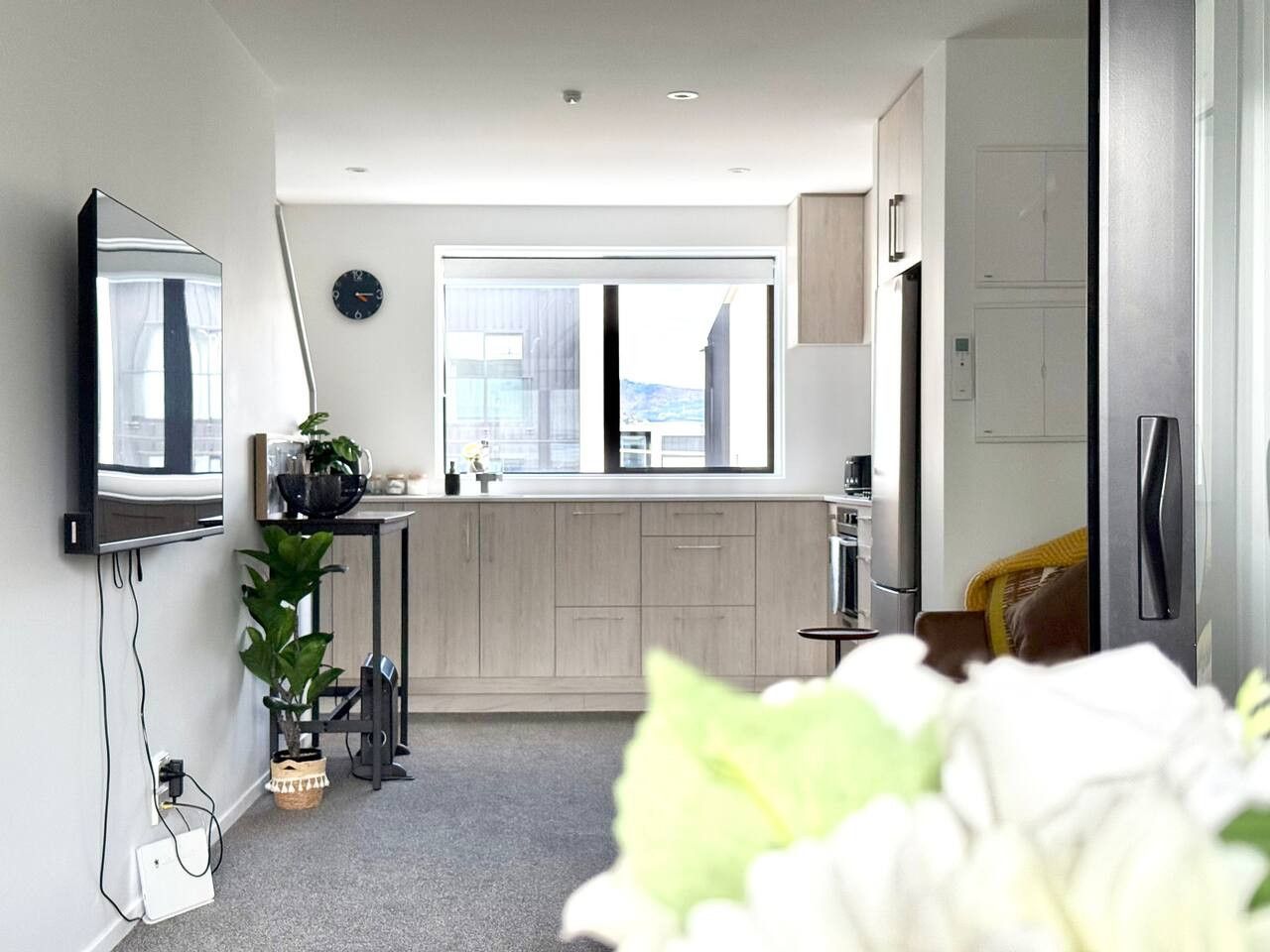 A modern, compact apartment with a kitchenette, a large window, and a flat-screen TV mounted on the wall.