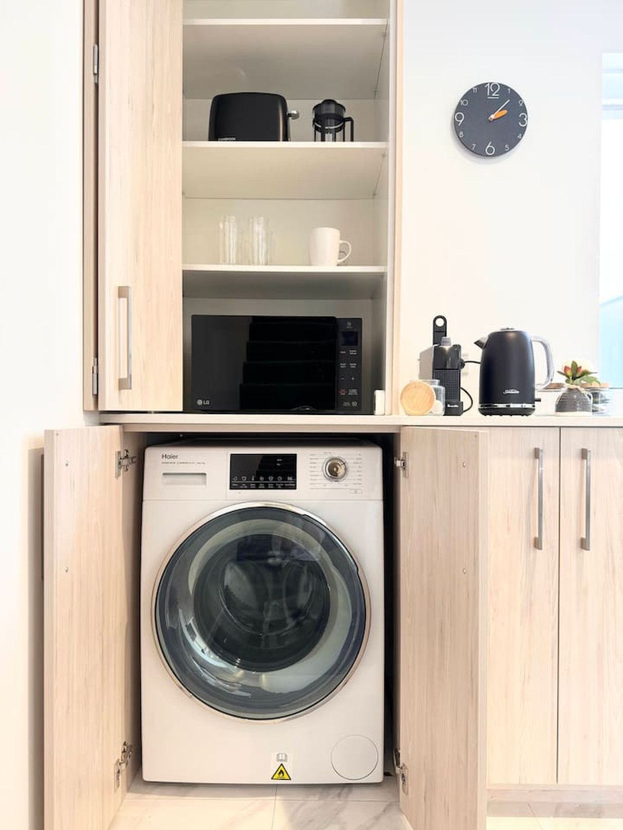 A compact laundry area with a front-loading washer and dryer, a coffee maker, and a clock on the wall.
