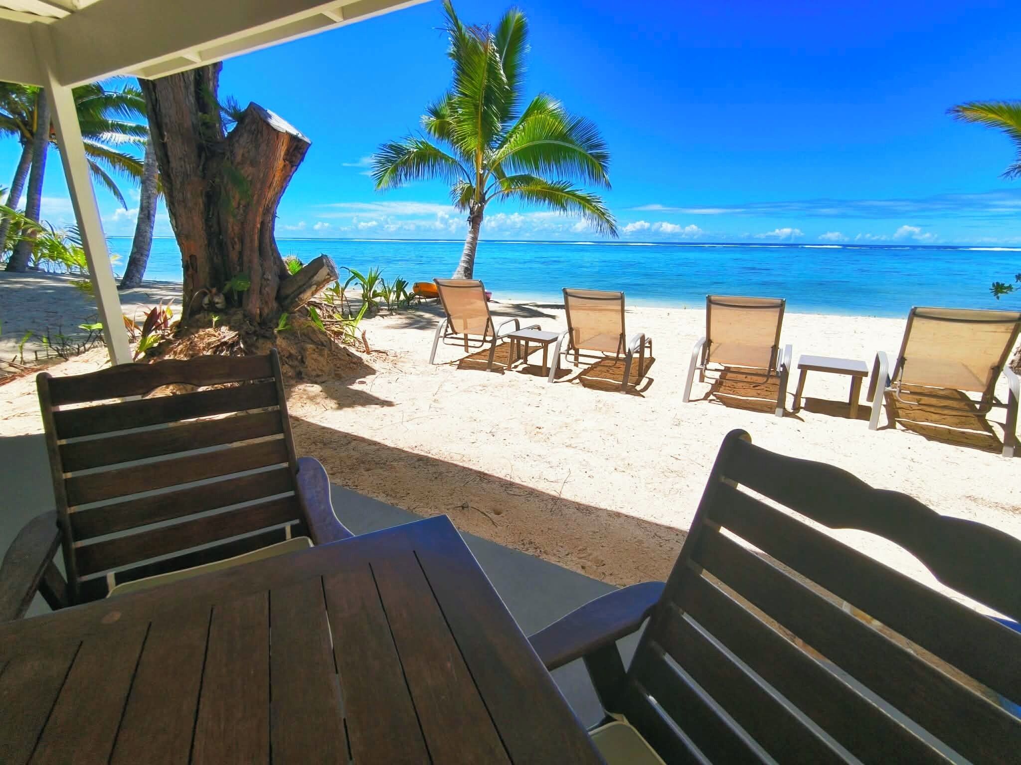 A beachside seating area with wooden tables and chairs, a palm tree, and a view of the ocean.