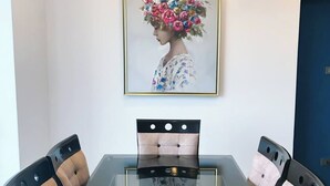 Ett matbord med en glasskiva och fyra stoppade stolar. En inramad tavla med ett blomsterarrangemang hänger på väggen ovanför bordet.