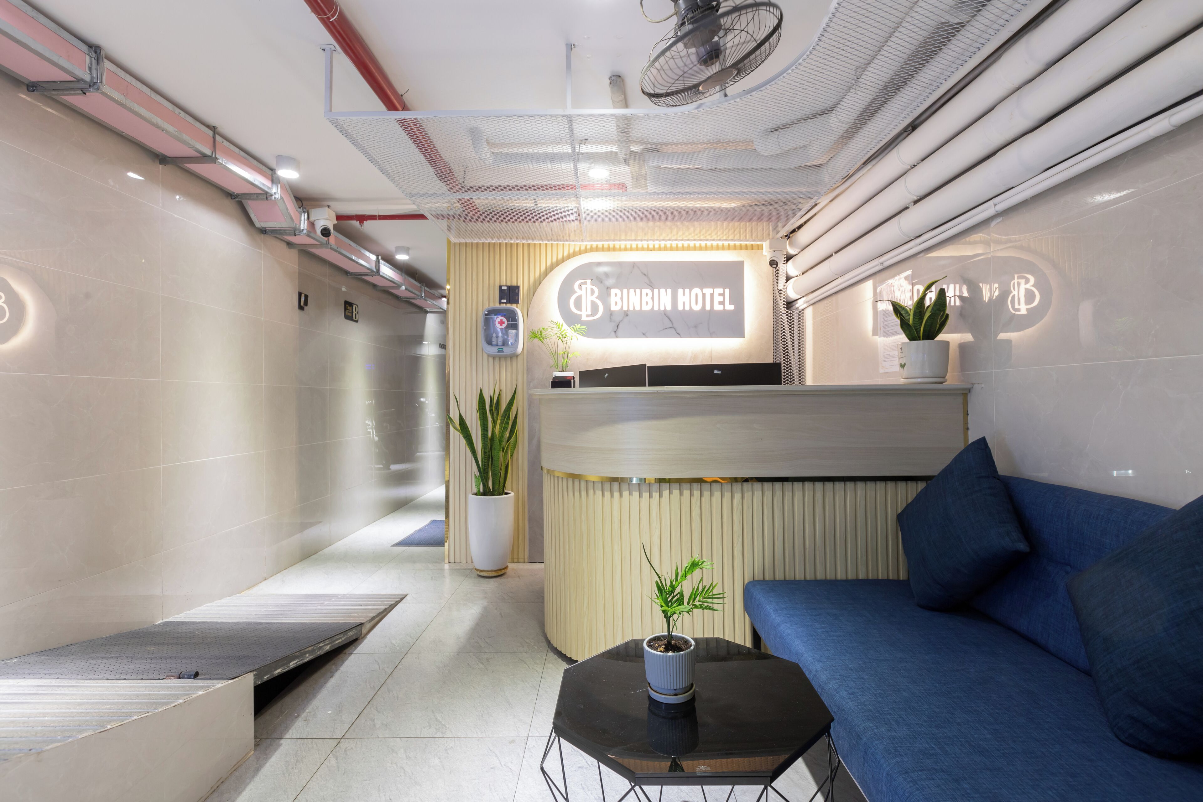 Ein Hotel-Empfangsbereich mit einer Rezeption, einem blauen Sofa, einem kleinen Tisch mit Pflanze und einem Blumentopf an der Wand.