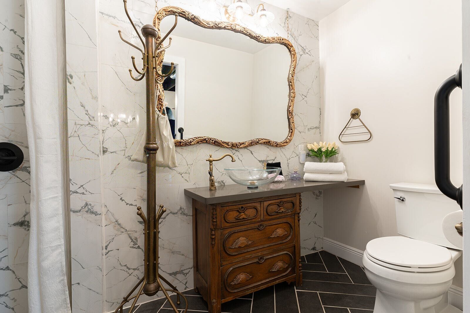 A bathroom with a marble wall, a wooden vanity with drawers, a glass bowl sink, a white toilet, and a gold-framed mirror.