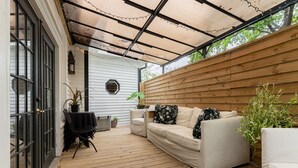 A covered patio area with a wooden deck, a white sofa with floral cushions, and potted plants. - The August Inn (Spring)