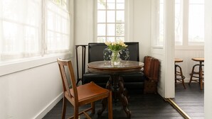 A room with a round wooden table, a black upholstered chair, a wooden chair, and a vase with flowers. - The August Inn (Spring)
