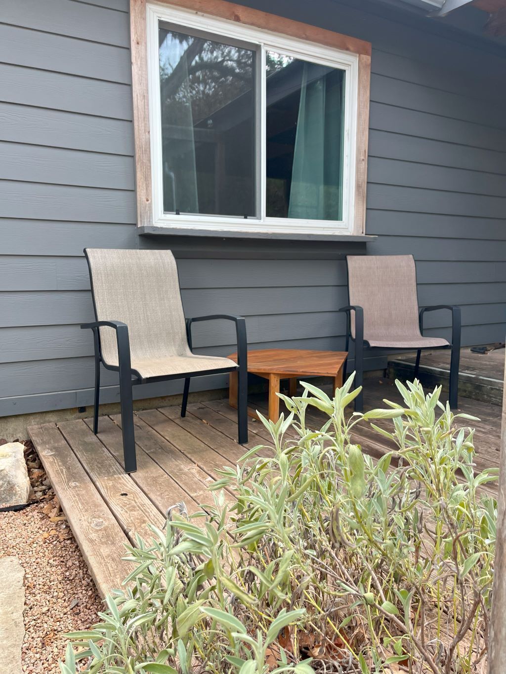 Een houten terras met twee stoelen en een tafel, een raam met gordijnen, en een plant op de voorgrond.