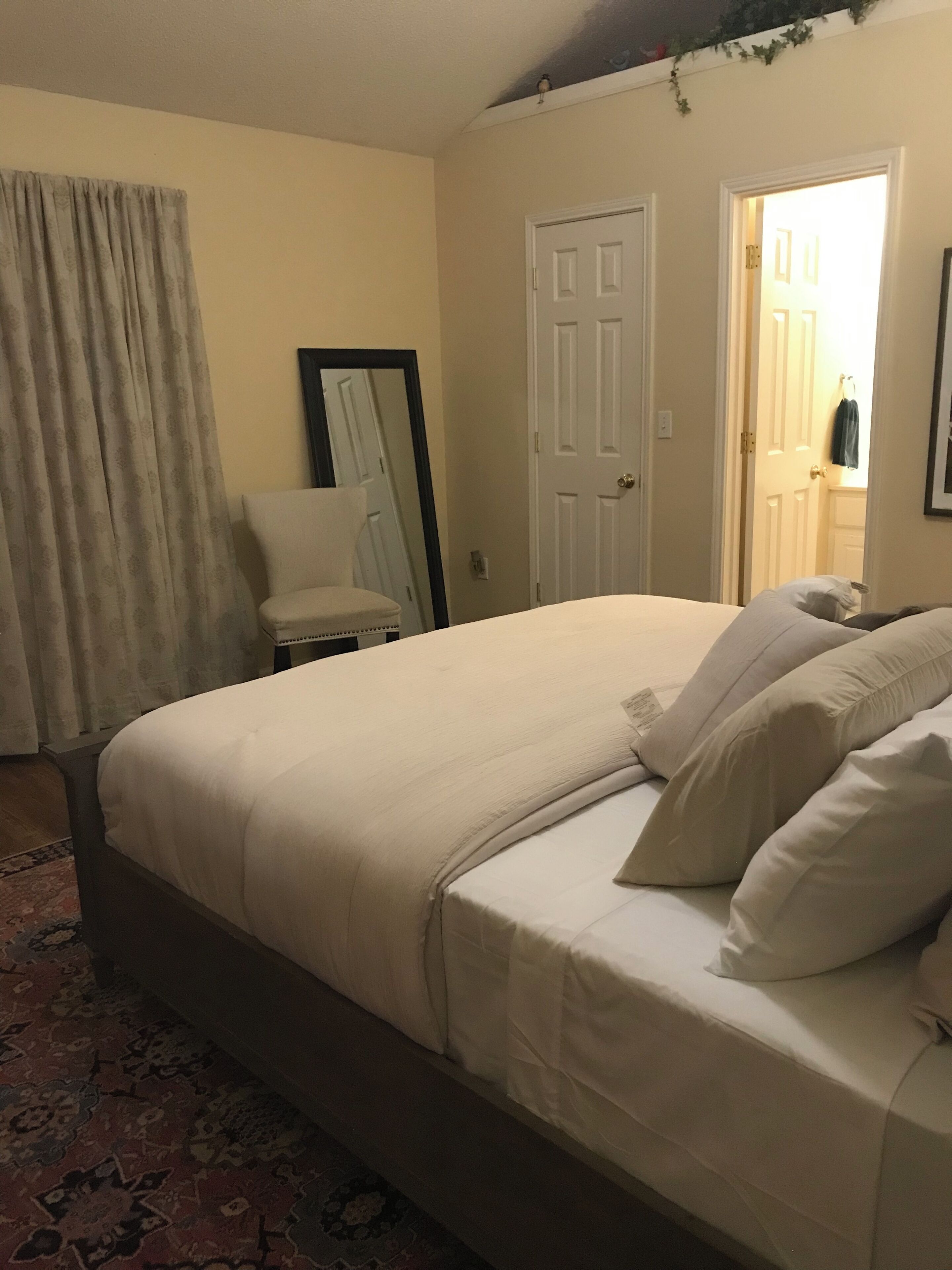 Une chambre bien rangée, avec un grand lit, une chaise et une porte donnant sur une autre pièce.