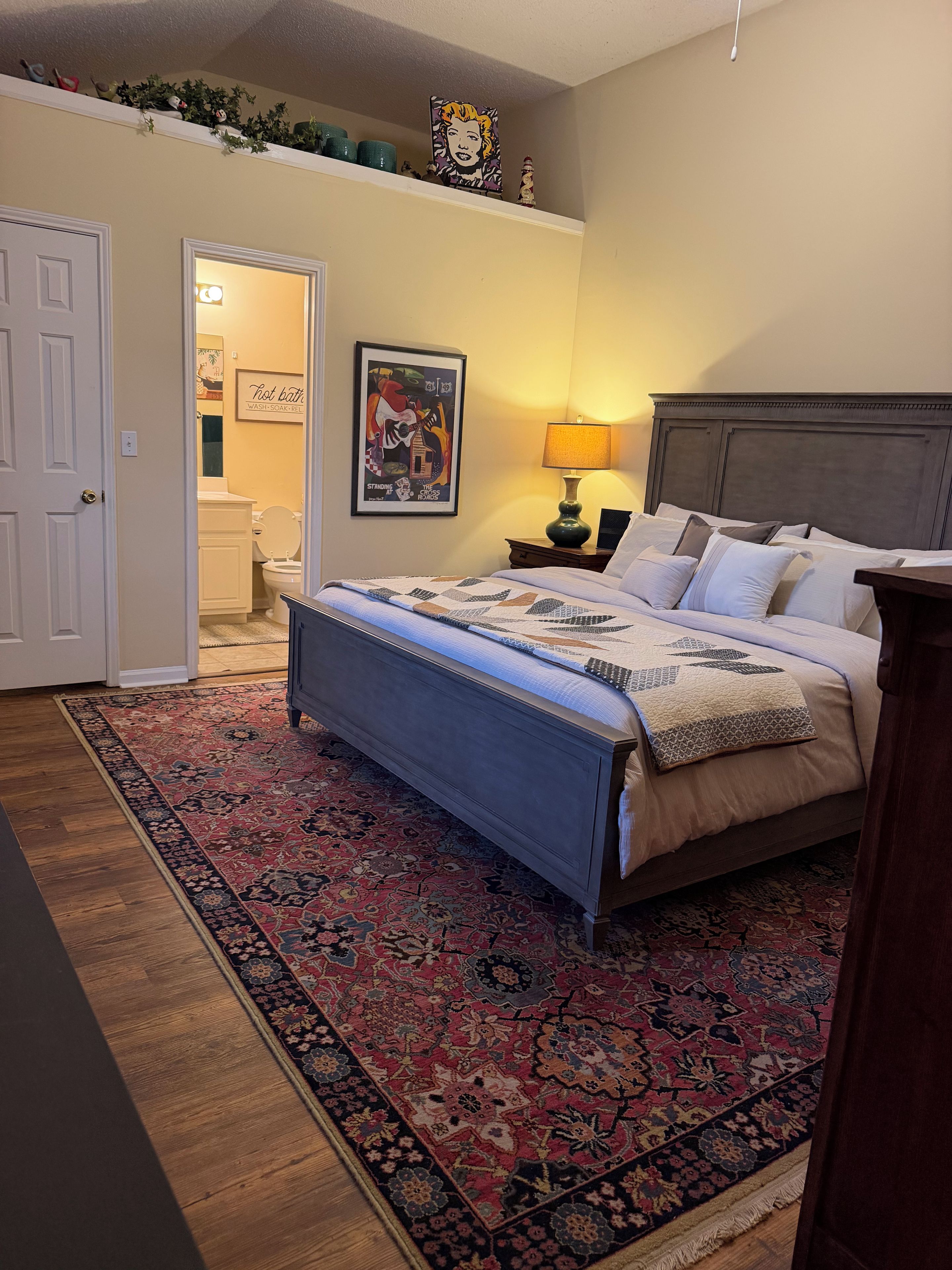 A bedroom with a bed, a nightstand, a lamp, a rug, and a door leading to a bathroom.