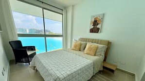 A bedroom with a large bed, a chair, a view of the ocean, and a painting on the wall. - Cartagena Baia Kristal | Perfect Escape Kabo 1-Apt 2 F (Cartagena de Indias)