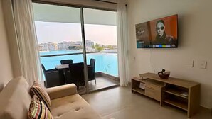 A living room with a sofa, a TV mounted on the wall, a wooden TV stand, and a sliding glass door leading to a balcony with a pool view. - Cartagena Baia Kristal | Perfect Escape Kabo 1-Apt 2 F (Cartagena de Indias)