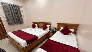 A hotel room with two wooden beds, each with white and red bedding, a wooden nightstand, and a grey patterned curtain. - Hotel Hare Krishna Dham (Mathura)