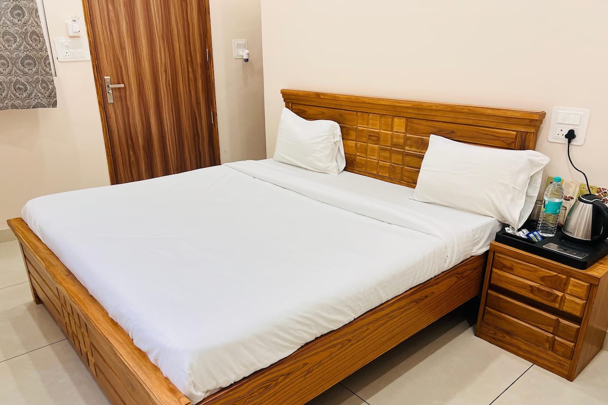 A neatly made bed with white linens, a wooden headboard, and a bedside table with a water bottle, a kettle, and a power strip.