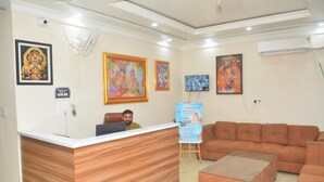 A reception area with a wooden reception desk, a brown sofa, and a coffee table. There are framed pictures on the walls and a poster for Jay Vidhavan Travels. - Hotel Hare Krishna Dham (Mathura)