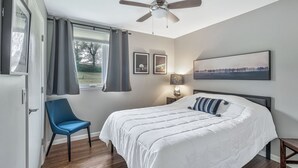 Une chambre avec un grand lit, une chaise, un ventilateur de plafond, une fenêtre avec des rideaux et des tableaux encadrés accrochés au mur.