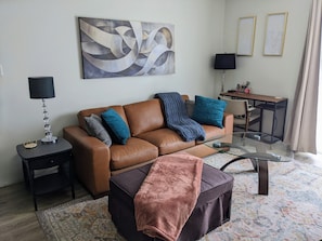 Uma sala de estar com um sofá de couro marrom, uma mesa de centro de vidro, um tapete estampado, uma mesa lateral com abajur e um quadro na parede.