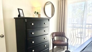 A black dresser with drawers, a mirror, and a small plant on top. There is a brown chair next to the dresser and a window with curtains. - Steps from Shands. Ready for Rotations: Mid-July and October Rotations Available (Gainesville)