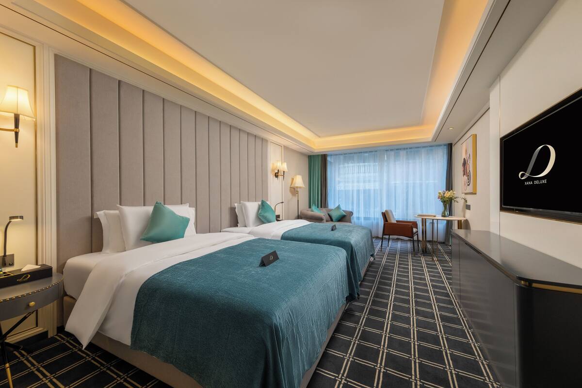 A hotel room with two beds, a desk, a TV, and a window with curtains.
