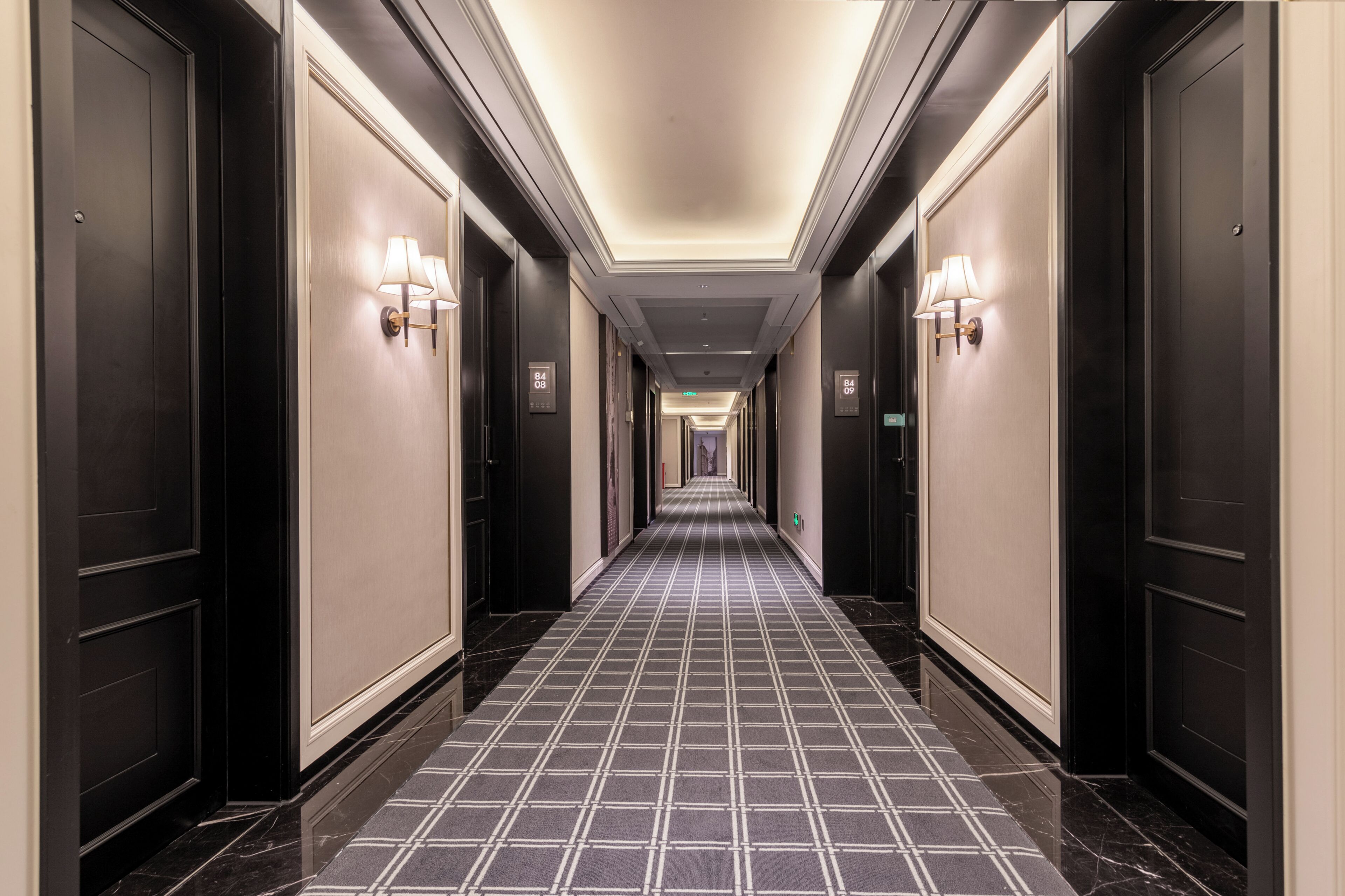 A long, narrow hotel corridor with dark doors on either side, light wall sconces, and a checkered floor pattern.