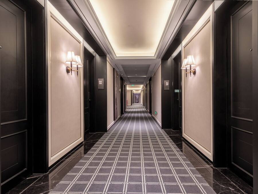 A long, narrow hotel corridor with dark doors on either side, light wall sconces, and a checkered floor pattern.